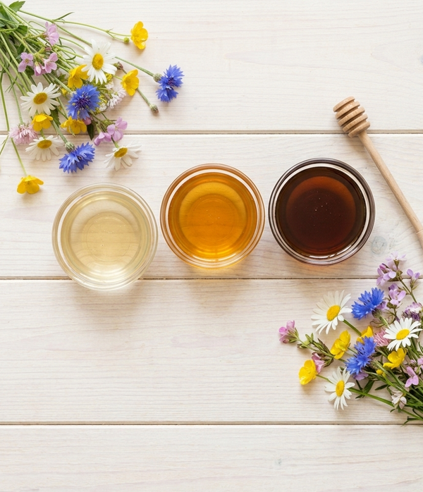 Three glass jars of honey in different shades — light, golden and dark — on a pale wooden table, with assorted wildflowers in two corners